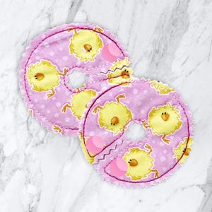 May include: Two pink fabric nursing pads with a scalloped edge and a yellow chick pattern. The pads have a circular shape with a center opening and a zig-zag seam. The background is a white marble surface.