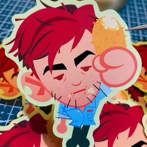 Könnte beinhalten: Ein gestanzter Aufkleber mit einer Cartoonfigur mit roten Haaren, Sommersprossen und einem blauen Hemd, die einen Corn Dog hält. Der Aufkleber hat einen hellgrünen Rand. Die Buchstaben "SDN" sind auf dem Hemd zu sehen.