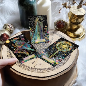 May include: Three tarot cards on a wooden surface. The cards are "The Empress", "The Fool", and "Wheel of Fortune". Each card has a black background with colourful illustrations and gold accents.