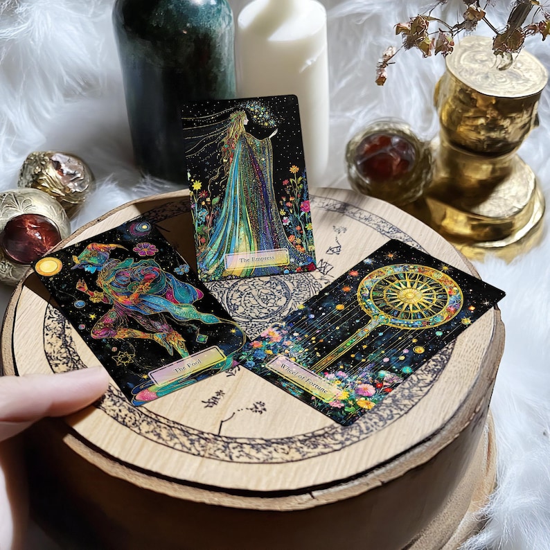 May include: Three tarot cards on a wooden surface. The cards are "The Empress", "The Fool", and "Wheel of Fortune". Each card has a black background with colourful illustrations and gold accents.
