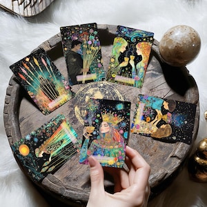 May include: A set of tarot cards with gold foil accents and colourful illustrations. The cards feature images of people, flowers, and other symbols. The cards are laid out on a wooden surface.