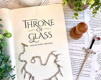 May include: A silver necklace with a red gemstone charm and silver spikes. The necklace is laying on a book titled "Throne of Glass: Collector's Edition".