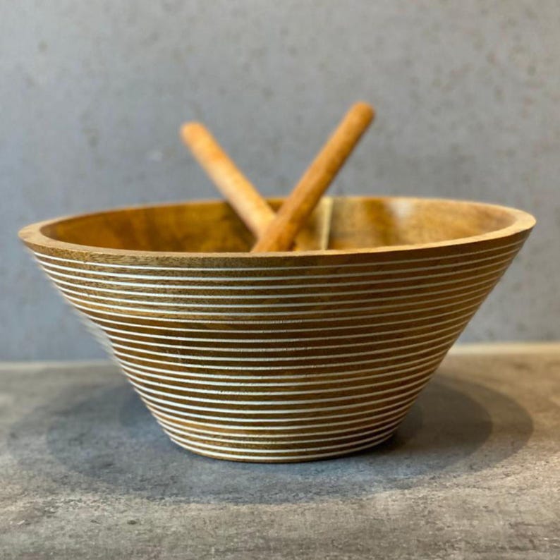May include: A wooden salad bowl with a wide, flared design. The bowl is light brown with horizontal white stripes. Two wooden salad serving spoons are inside the bowl. The bowl is on a gray surface.
