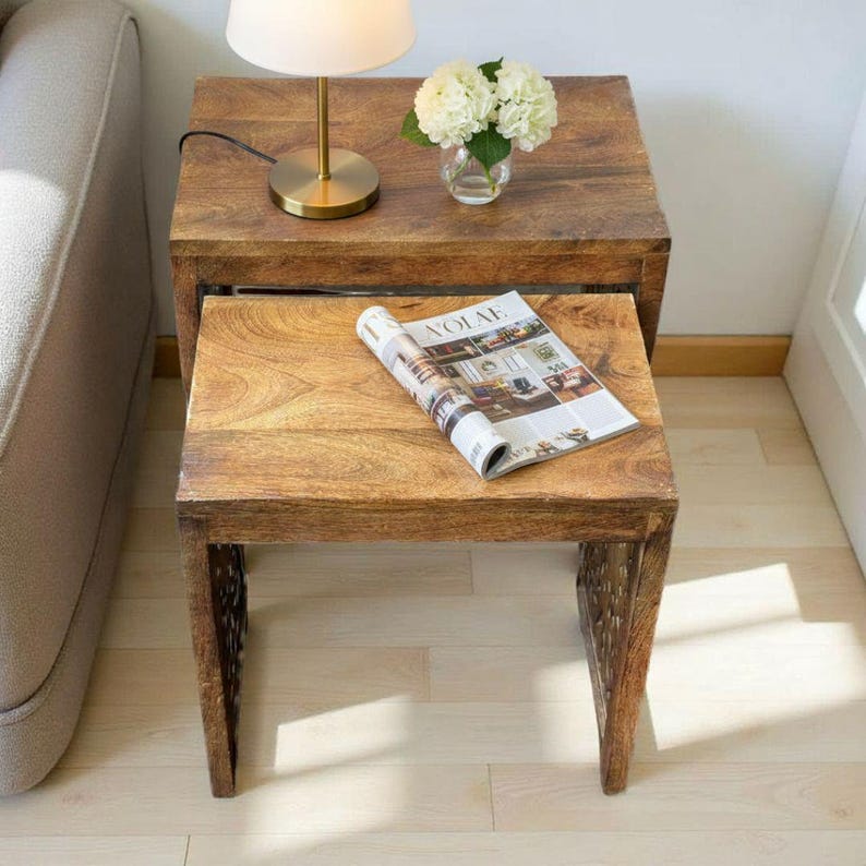 Pode incluir: Duas mesas de encaixe de madeira com acabamento natural. A mesa superior tem uma pequena l&acirc;mpada com base dourada e um vaso com flores brancas. A mesa inferior tem uma revista aberta. As mesas s&atilde;o de cor castanha quente.
