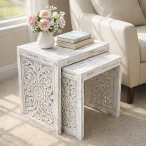 May include: Two white nesting tables with carved floral designs. The tables are topped with books and a white vase holding pink and white flowers. The tables are made of wood and have a distressed finish, creating a rustic look.