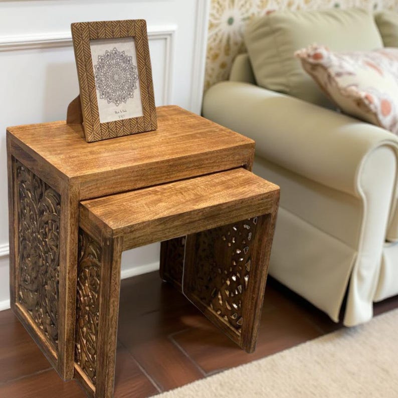 Pode incluir: Duas mesas de encaixe de madeira castanha quente, com entalhes florais intrincados nas laterais. As mesas t&ecirc;m um acabamento polido. Uma foto emoldurada com um design de mandala est&aacute; em cima da mesa maior.
