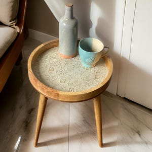 May include: A small wooden side table with three legs and a round top. The top is decorated with a white and light blue floral pattern. A blue and white ceramic mug and a grey ceramic bottle are on the table.