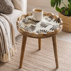 May include: A round wooden side table with three tapered legs and a scalloped edge. The tabletop features a white surface with a black botanical leaf pattern. A white ceramic mug filled with coffee sits on the center of the table surface.