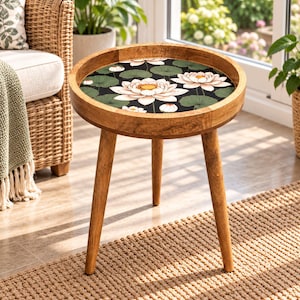 May include: A round wooden side table with three tapered legs. The tabletop features a dark background with a printed pattern of white and pink lotus flowers and green lily pads. The wood has a natural light brown finish.