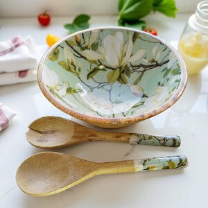以下が含まれることがあります： 花柄のデザインの木製サラダボウルと2つの木製サラダスプーン。ボウルは、白いモクレンの花と茶色の縁が付いた、明るい緑色の内側です。スプーンは、ハンドルに花柄のデザインが施された明るい木の色をしています。