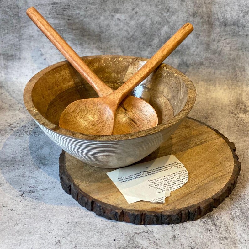 May include: A wooden salad bowl with two matching wooden salad spoons. The bowl is light brown with a white stripe design around the outside. The spoons are crossed inside the bowl. The bowl sits on a wooden round serving board.
