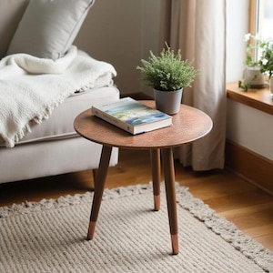 Pode incluir: Uma pequena mesa lateral redonda com tampo cor de cobre e três pernas de madeira. Um livro e uma planta em vaso estão sobre a mesa. A mesa está sobre um tapete neutro, perto de uma janela e de um sofá.