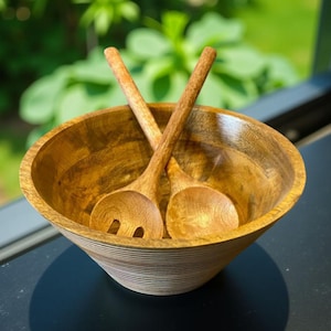 May include: A wooden salad bowl with two matching wooden salad servers. The bowl is round with a wide opening and a textured, ridged exterior. The salad servers are crossed over the bowl.