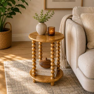 Op de afbeelding: Een ronde, lichtbruine houten bijzettafel met een kralenpootontwerp. De tafel heeft een bovenste plank en drie kleine voetjes. Een witte vaas met groen, een kaars, een houten kom en boeken staan op de tafel.