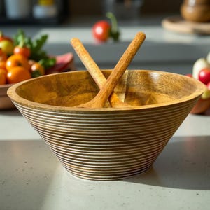 May include: Wooden salad bowl with two wooden salad servers. The bowl is light brown with white horizontal stripes around the exterior. The salad servers are crossed inside the bowl. The background includes tomatoes and other produce.