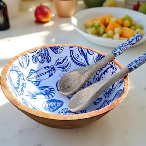 Könnte beinhalten: Eine Holzsalatschüssel mit weißem Innenraum und blauen Illustrationen von Meereskreaturen. Zwei hölzerne Salatbestecke mit blau gemusterten Griffen liegen in der Schüssel. Die Schüssel hat einen natürlichen Holzrand.