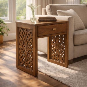 May include: A brown wooden side table with a drawer and carved floral patterns. The table is about 61 cm tall. A small glass vase with flowers and books are on top. The table is placed next to a beige sofa.