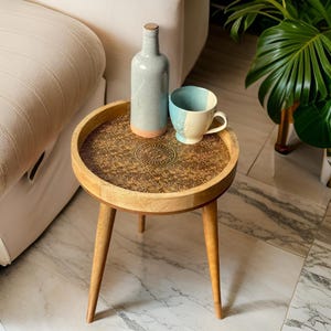 Może przedstawiać: Mały drewniany stolik pomocniczy z trzema nogami i okrągłym blatem. Blat ma ozdobny wzór i jest wykonany z drewna. Na stoliku stoi niebieska i biała ceramiczna filiżanka oraz niebieska ceramiczna butelka.