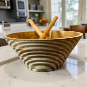 May include: A wooden salad bowl with a wide, flared design and horizontal white stripes. Two wooden salad servers rest inside the bowl. The bowl is made of light brown wood and sits on a white countertop.
