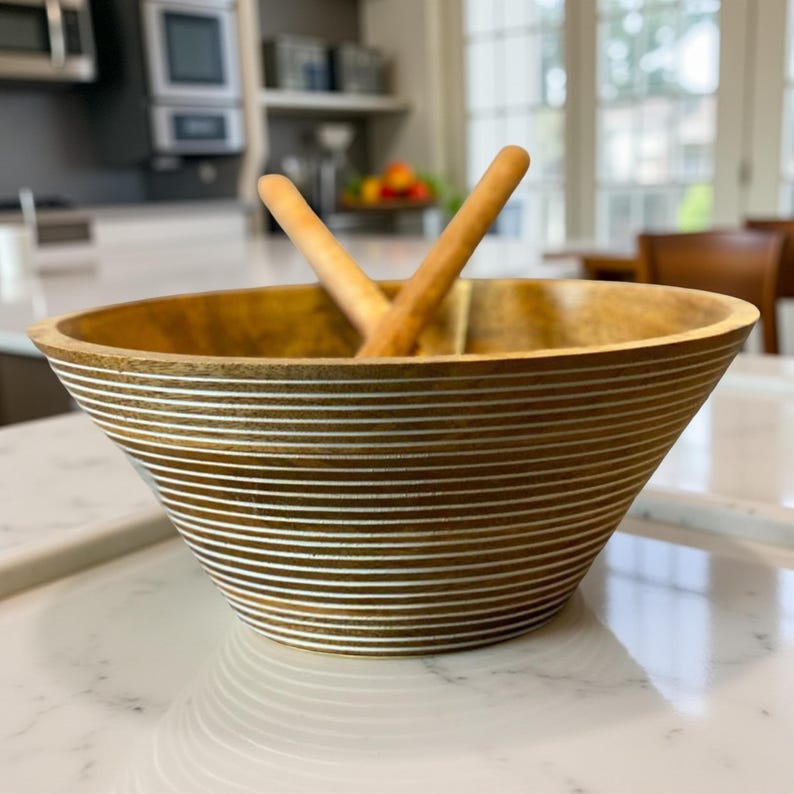 May include: A wooden salad bowl with a wide, flared design and horizontal white stripes. Two wooden salad servers rest inside the bowl. The bowl is made of light brown wood and sits on a white countertop.