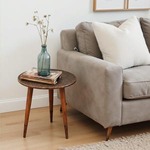 May include: A small, round side table with a dark, textured tabletop and three wooden legs. A blue glass bottle with dried flowers and a book rest on the table. A grey sofa with a white pillow is in the background.