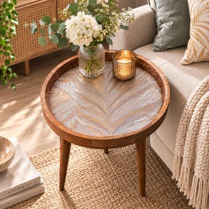 May include: A round wooden side table with three tapered legs and a tabletop featuring a carved, light-coloured leaf pattern. On the table sits a clear, ribbed glass vase filled with white hydrangea flowers and a small, gold-toned glass candle holder.