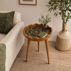 May include: A round wooden side table with three tapered legs and a scalloped edge. The tabletop features a black surface with a gold-colored floral design. A small glass vase with green sprigs and a closed book rest on the table surface.