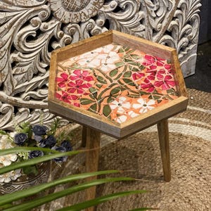 Könnte beinhalten: Kleiner Beistelltisch aus Holz mit sechseckiger Tischplatte. Die Tischplatte hat ein Mosaik-Blumenmuster in Rosa und Weiß auf orangefarbenem Grund. Der Tisch steht auf drei Holzbeinen, ideal für die Inneneinrichtung.