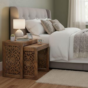Pode incluir: Uma cena de quarto com uma cama com cabeceira estofada cinza e roupa de cama branca. Duas mesas de encaixe de madeira com desenhos florais esculpidos est&atilde;o ao lado da cama. Uma l&acirc;mpada branca, uma pequena suculenta e uma caneca est&atilde;o nas mesas.