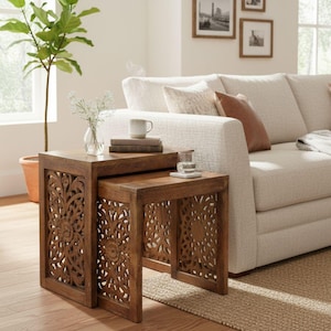 May include: Two nesting wooden side tables with intricate floral cutouts. The tables are a warm brown color and have a square shape. A small glass of water and a mug sit on top of the tables. The tables are next to a beige sofa.