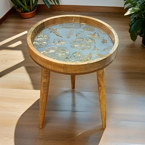 Może przedstawiać: Okrągły drewniany stolik pomocniczy z trzema nogami. Blat stołu ma wzór kwiatowy w kolorze niebieskim i złotym.