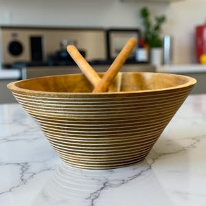 May include: A wooden salad bowl with a wide, flared shape. The bowl is light brown with horizontal white stripes. Two wooden salad serving utensils are inside the bowl. The bowl is on a white marble countertop.