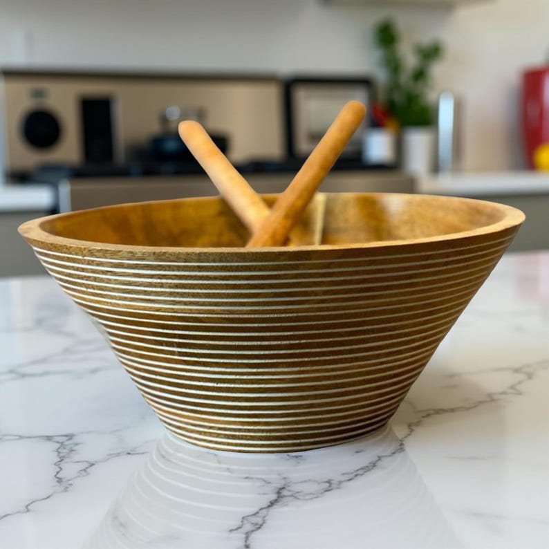 May include: A wooden salad bowl with a wide, flared shape. The bowl is light brown with horizontal white stripes. Two wooden salad serving utensils are inside the bowl. The bowl is on a white marble countertop.