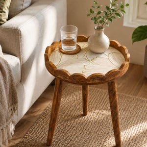 May include: A small, round wooden side table with a scalloped edge. The tabletop has a cream surface with a gold floral design. A glass of water, a coaster, and a small vase with greenery are on the table. The table has three wooden legs.