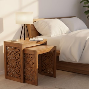 Pode incluir: Duas mesas de encaixe de madeira com entalhes florais intrincados. Uma pequena l&acirc;mpada com uma sombra branca est&aacute; na mesa maior, junto com um livro e &oacute;culos. As mesas est&atilde;o em um quarto, ao lado de uma cama com roupa de cama branca.