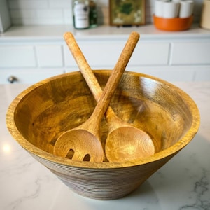 May include: A wooden salad bowl with two matching wooden salad servers. The bowl is round with a wide opening and a textured, striped exterior. The salad servers are crossed inside the bowl, ready for use.
