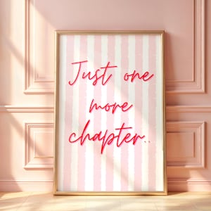 May include: A framed print with the text "Just one more chapter" in red script. The background features vertical pink and white stripes. The frame is gold, and the print is leaning against a pink wall.