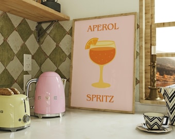 May include: A kitchen scene featuring a pink Smeg kettle and a yellow toaster. A framed poster with an Aperol Spritz design is propped up on the counter. A black and white patterned cup and saucer sit nearby.