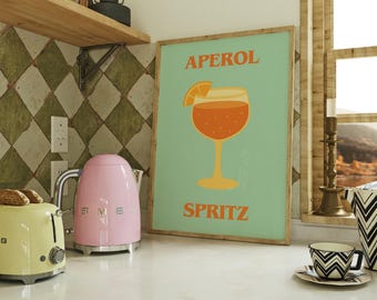 May include: A framed poster with the words "APEROL SPRITZ" and an illustration of the cocktail. A pink electric kettle and a yellow toaster sit on the counter. A black and white patterned cup and saucer are also on the counter.