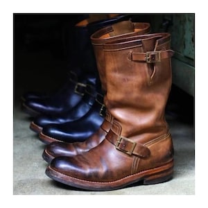 May include: A pair of mid-calf leather engineer boots in a distressed brown finish, featuring a rounded toe, a stacked heel, and two adjustable straps with brass-toned metal buckles located at the ankle and the upper shaft of the boot.