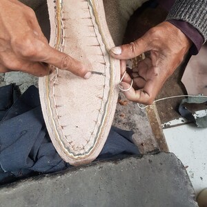 Op de afbeelding: Close-up van schoenmaken. Een lichtbruine leren schoenzool wordt met wit garen met de hand gestikt. De zool heeft een lichtbruin interieur en een donkerdere bruine rand. De handen van de schoenmaker zijn zichtbaar, die de naald vasthouden en de draad geleiden.