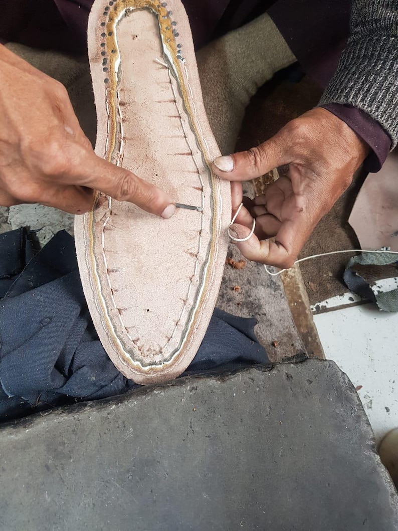 Op de afbeelding: Close-up van schoenmaken. Een lichtbruine leren schoenzool wordt met wit garen met de hand gestikt. De zool heeft een lichtbruin interieur en een donkerdere bruine rand. De handen van de schoenmaker zijn zichtbaar, die de naald vasthouden en de draad geleiden.
