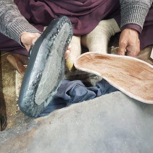 Op de afbeelding: Een schoenmaker houdt een zwarte schoenzool en een houten binnenzool vast. De schoenzool is zwart met een textuuroppervlak en zichtbare stiksels. De binnenzool heeft een lichtbruine kleur.
