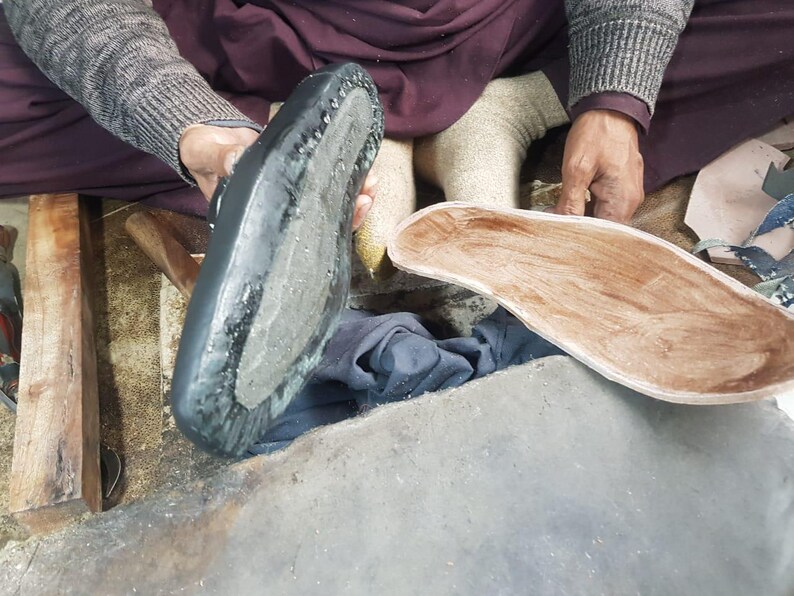 Op de afbeelding: Een schoenmaker houdt een zwarte schoenzool en een houten binnenzool vast. De schoenzool is zwart met een textuuroppervlak en zichtbare stiksels. De binnenzool heeft een lichtbruine kleur.
