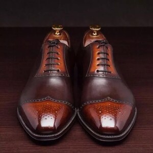 May include: A pair of brown leather lace-up shoes with a cap toe design. The toe is a lighter shade of brown with a brogue pattern.