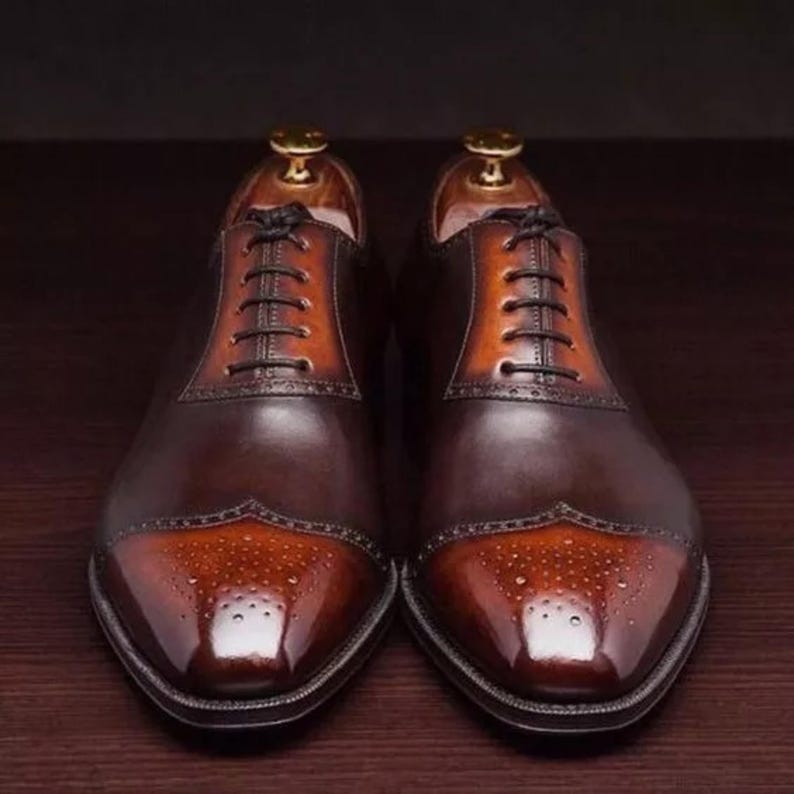 May include: A pair of brown leather lace-up shoes with a cap toe design. The toe is a lighter shade of brown with a brogue pattern.