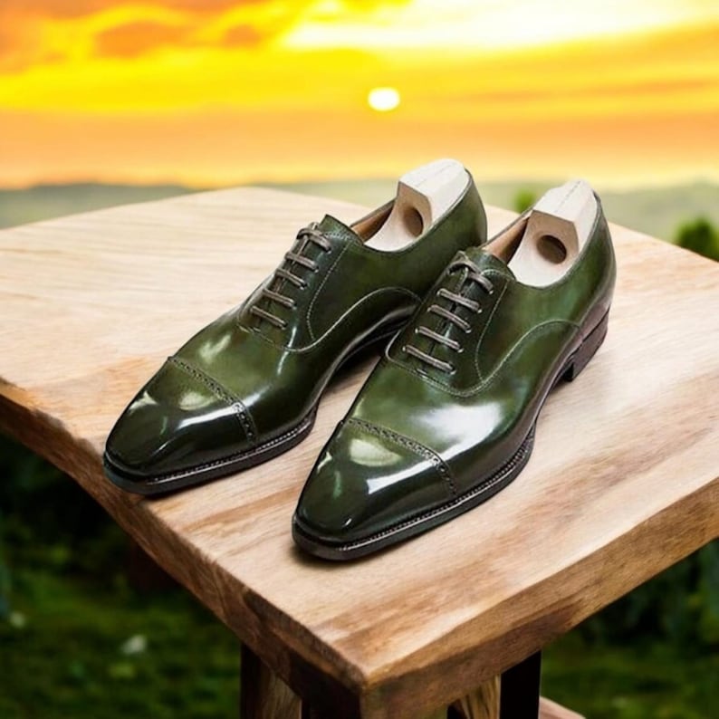 Op de afbeelding: Een paar groene leren herenschoenen met een glanzende afwerking. De schoenen hebben een klassiek Oxford-ontwerp met veters en een neuskap. Houten schoenspanners zijn erin geplaatst. De schoenen worden getoond op een houten oppervlak met een vage achtergrond.
