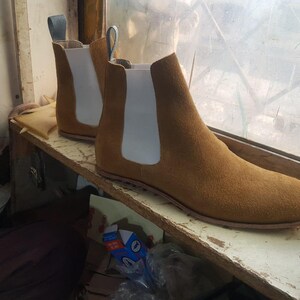 Op de afbeelding: Een paar bruin su&egrave;de Chelsea boots met witte elastische zijpanelen en treklussen. De laarzen staan op een houten plank bij een raam, wat hun klassieke ontwerp benadrukt. Maat 42.