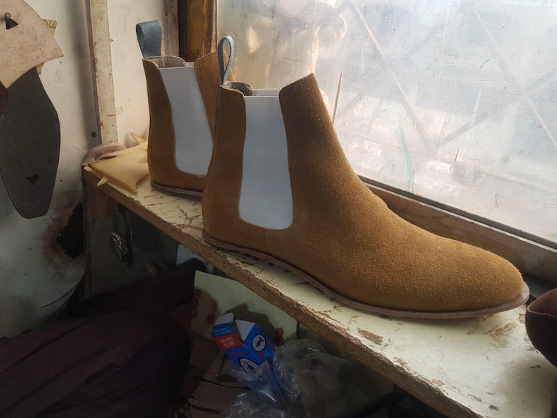 Op de afbeelding: Een paar bruin su&egrave;de Chelsea boots met witte elastische zijpanelen en treklussen. De laarzen staan op een houten plank bij een raam, wat hun klassieke ontwerp benadrukt. Maat 42.