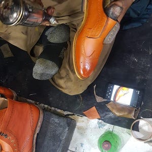 Op de afbeelding: Een schoenmaker werkt aan een paar oranje leren herenschoenen. De schoenen hebben decoratieve perforaties en een klassiek ontwerp. De schoenmaker gebruikt gereedschap om de schoen te vormen en te verfijnen, wat het vakmanschap laat zien.
